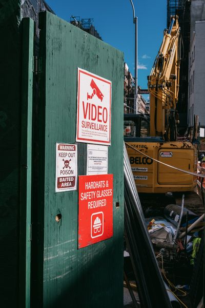 A partially open temporary green door covered with warning signs at a construction site, with an excavator visible in the background